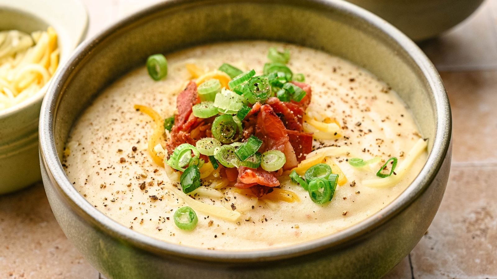 A bowl of creamy soup topped with chopped green onions, grated cheese, crispy bacon pieces, and ground black pepper.
