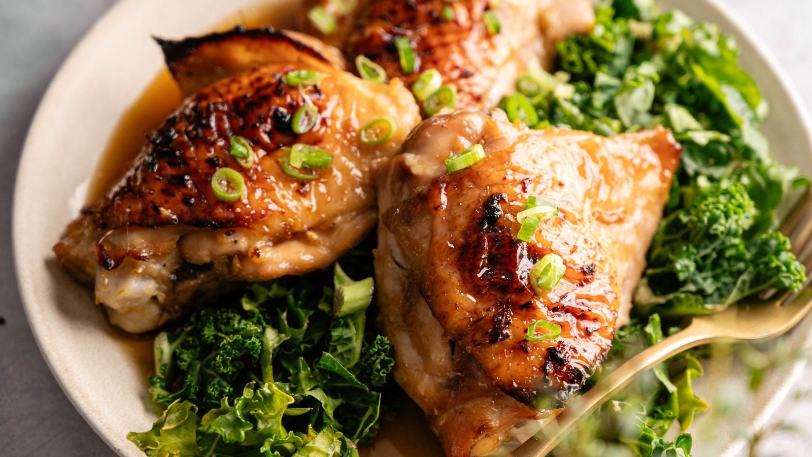 Three glazed chicken thighs served on a bed of leafy greens, garnished with chopped green onions, on a beige plate.