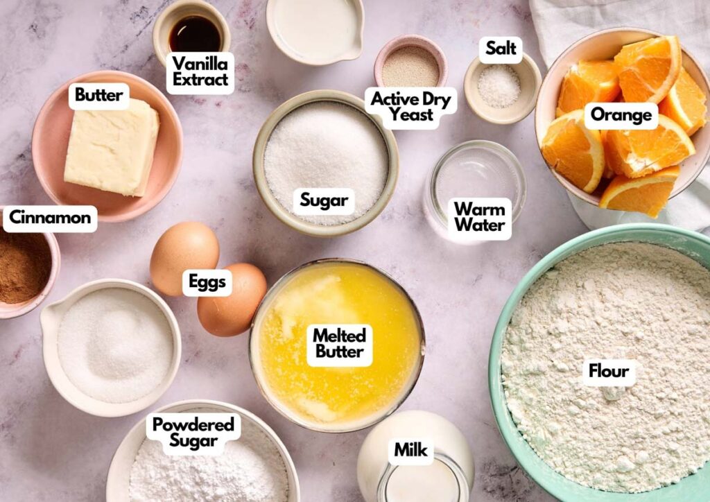 Various baking ingredients labeled in bowls and containers, including those needed for Air Fryer Orange Rolls: butter, eggs, sugar, flour, milk, orange slices, cinnamon, salt, yeast, powdered sugar, vanilla extract, and warm water.