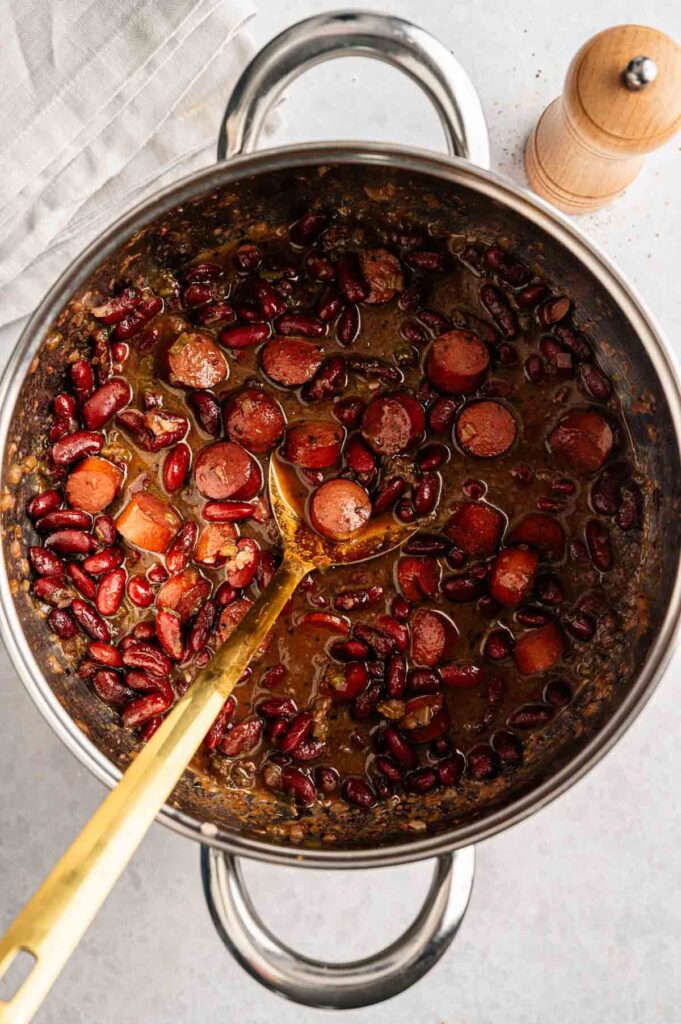 A metal pot filled with Red Beans and Rice, sliced sausage, and broth sits ready with a gold ladle inside. A wooden pepper grinder and a folded cloth napkin are nearby.