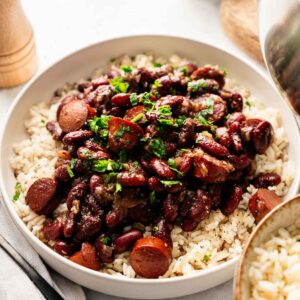 A plate of Red Beans and Rice features white rice topped with red beans, sliced sausage, and chopped herbs, served alongside a bowl of rice and a pepper grinder.