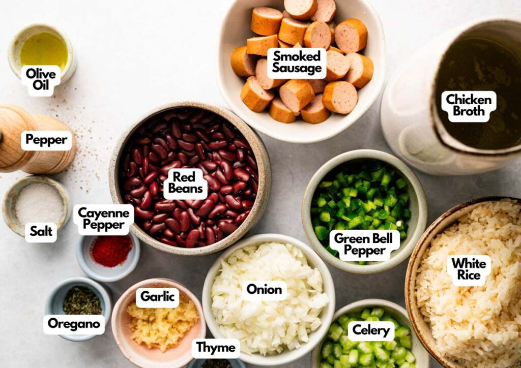 A collection of labeled ingredients in bowls for Red Beans and Rice, featuring smoked sausage, red beans, rice, green bell pepper, onion, celery, garlic, spices, chicken broth, olive oil, salt, and pepper.