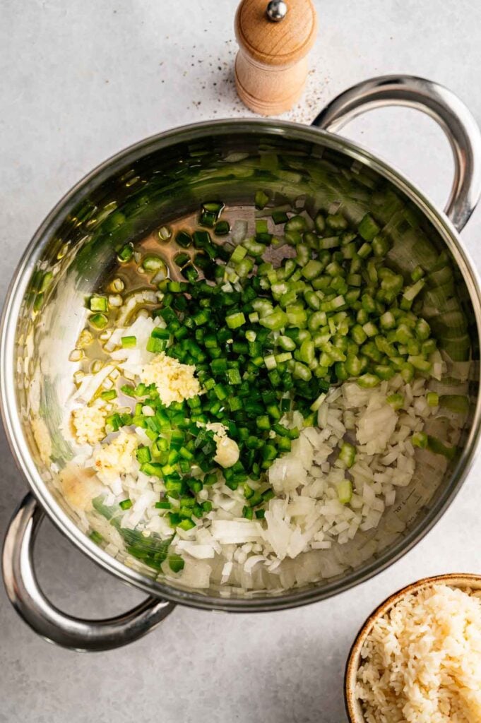 Chopped onions, celery, jalapeño, and minced garlic simmer in a stainless steel pot—the perfect base for classic Red Beans and Rice—with a pepper grinder and a bowl of cooked rice nearby on a light surface.