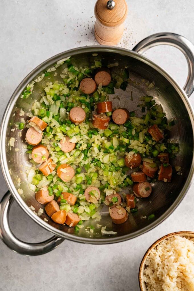 Chopped sausage, onions, and green bell peppers are sautéing in a stainless steel pot, the perfect base for a classic Red Beans and Rice dish. A pepper grinder and a bowl of rice sit nearby, ready to complete the meal.