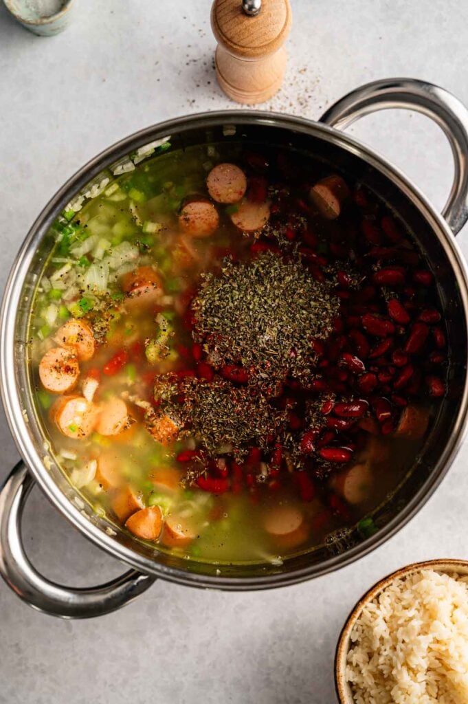 A stainless steel pot filled with sliced sausage, kidney beans, celery, spices, and broth captures the essence of classic Red Beans and Rice, resting next to a bowl of cooked white rice on a light countertop.