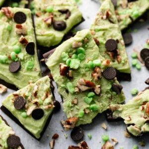 Shamrock Bark features pieces of green and dark chocolate bark topped with chocolate chips, chopped nuts, and green candy on a sheet of parchment paper.