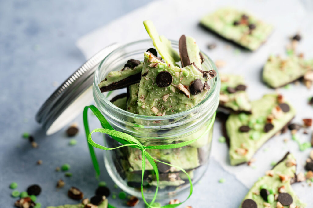 A glass jar filled with pieces of Shamrock Bark&mdash;green mint chocolate bark topped with chocolate chips and nuts&mdash;sits on a blue surface, with more delicious bark pieces scattered nearby.