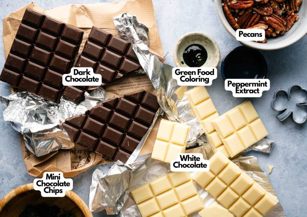 Bars of dark and white chocolate, mini chocolate chips, pecans, green food coloring, and peppermint extract arranged on a table with wrappers and a clover-shaped cookie cutter for making festive Shamrock Bark.