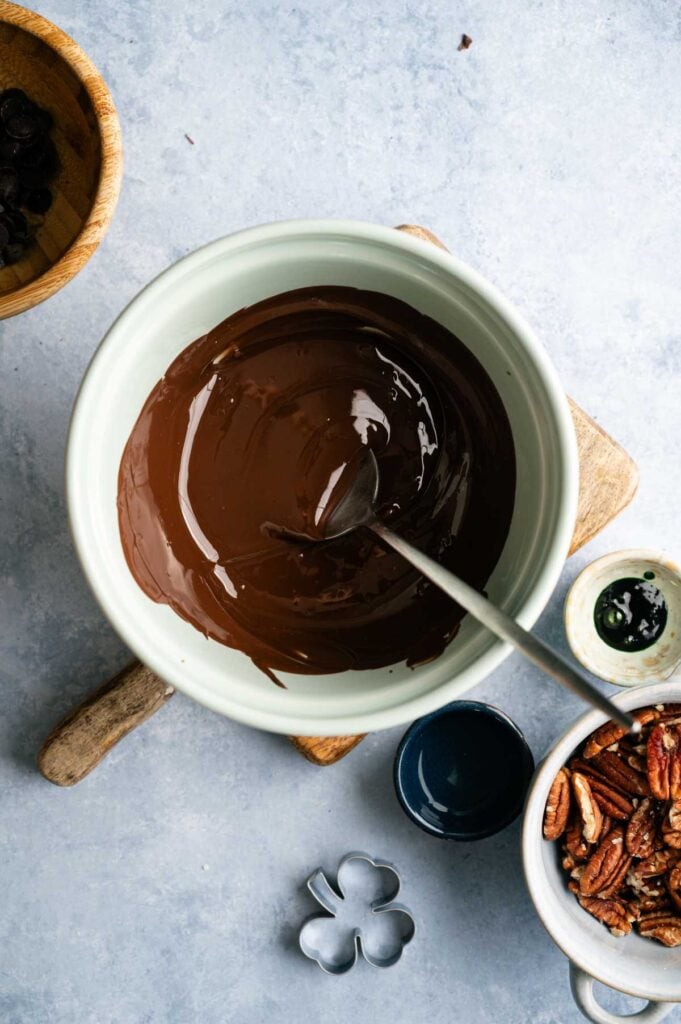 A bowl of melted chocolate with a spoon, surrounded by pecans, a wooden board, small bowls, and a shamrock-shaped cookie cutter on a light surface—perfect for creating festive Shamrock Bark.