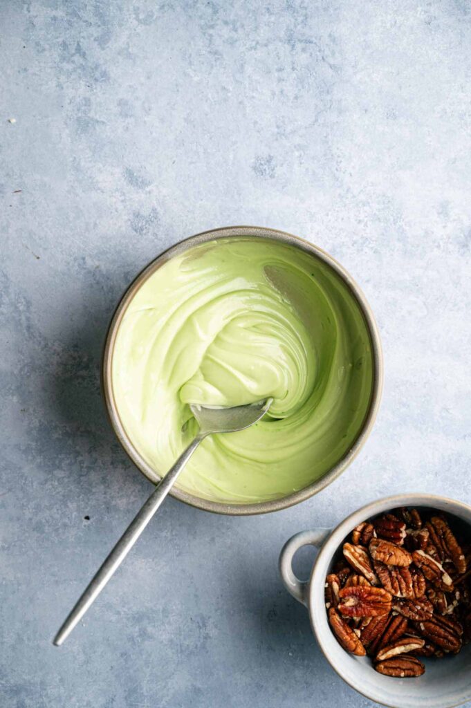 A bowl of green creamy mixture, reminiscent of the vibrant hues found in Shamrock Bark, sits with a spoon next to a smaller bowl filled with pecans, all set on a light blue textured surface.