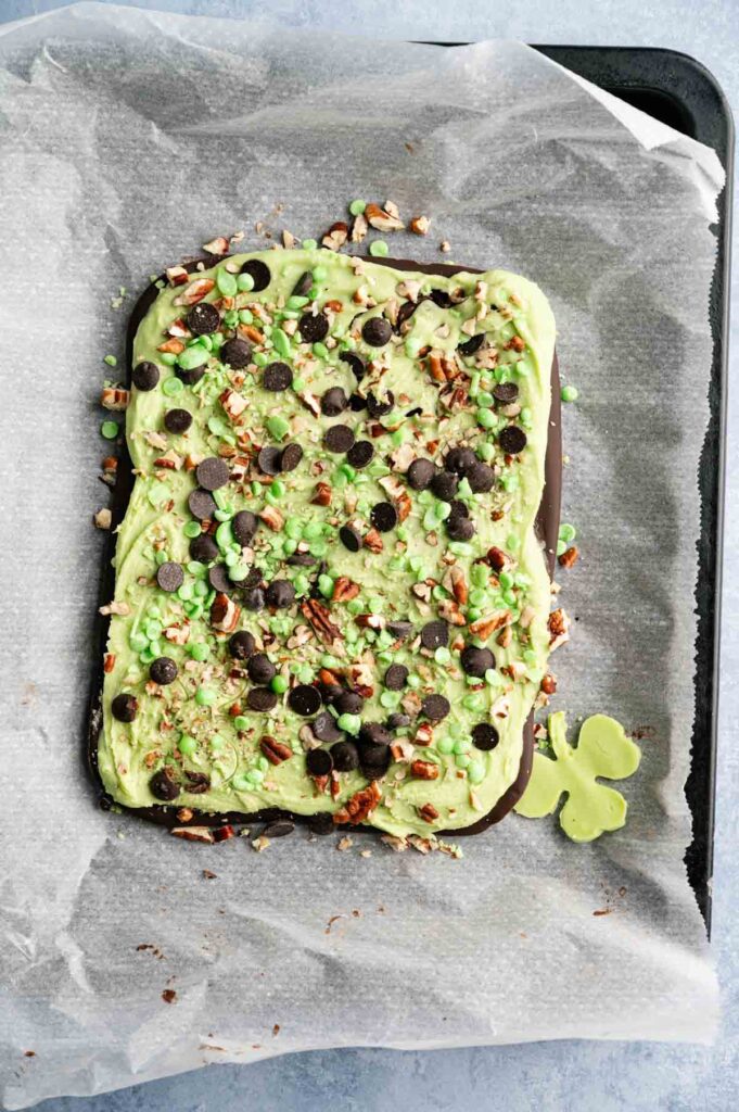A sheet of Shamrock Bark topped with green mint chocolate chips, dark chocolate chips, and chopped pecans on parchment paper.