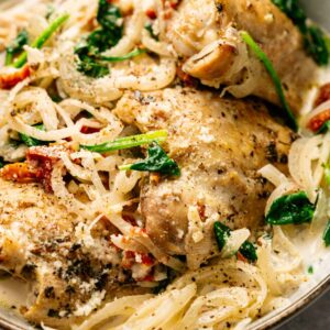 A bowl of Slow Cooker Chicken Thighs with saut&eacute;ed onions, spinach, sun-dried tomatoes, and creamy sauce, garnished with herbs.