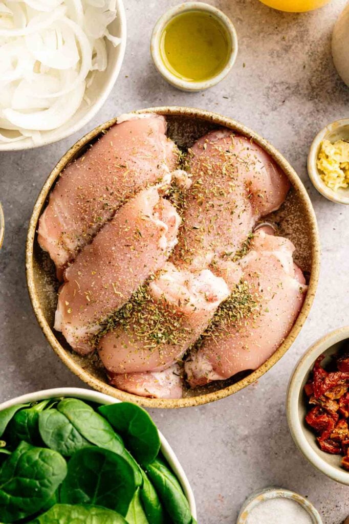 Raw chicken breasts seasoned with herbs in a bowl, surrounded by fresh spinach, sliced onions, minced garlic, olive oil, and sun-dried tomatoes on a gray surface—these same vibrant ingredients can also bring incredible flavor to Slow Cooker Chicken Thighs for an easy and delicious meal.