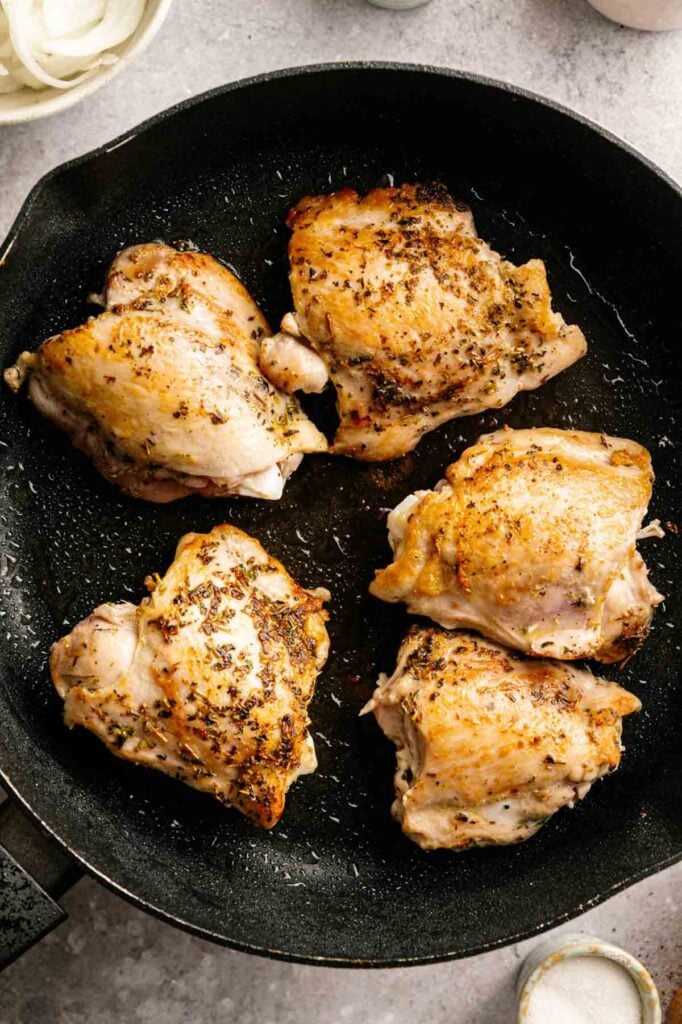 Four seasoned chicken thighs are cooking in a black skillet on a light gray countertop, with crispy, browned skin visible—perfect for making Slow Cooker Chicken Thighs that are tender and full of flavor.