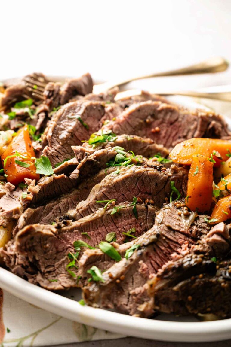 Sliced pot roast beef garnished with chopped herbs, served with cooked carrots on a white plate, reminiscent of a classic Slow Cooker Corned Beef with Cabbage meal.