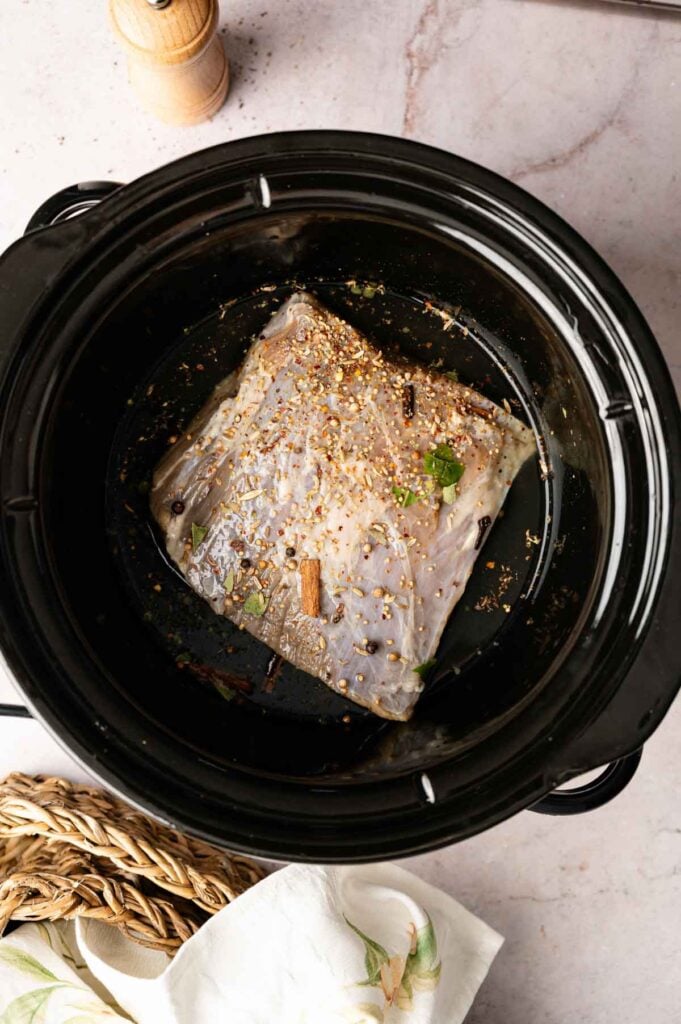Raw seasoned beef brisket in a slow cooker filled with water, ready to be cooked; spices and herbs are visible on the meat, making it perfect for a classic Slow Cooker Corned Beef with Cabbage.