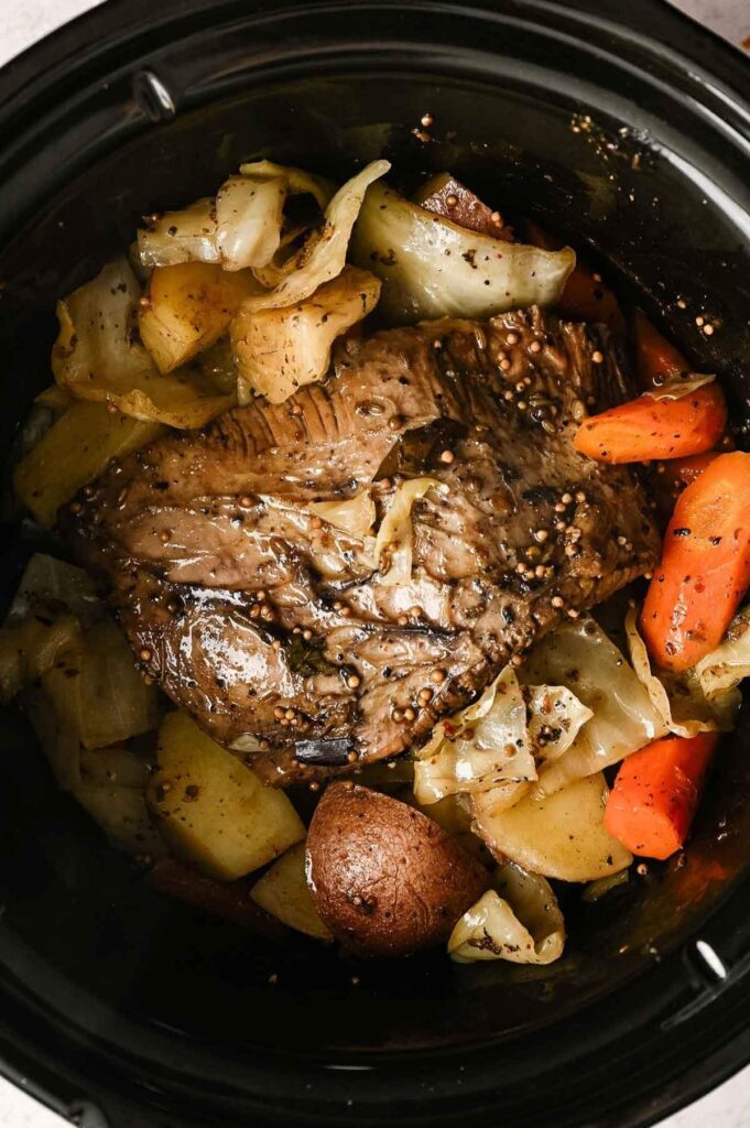 Overhead view of a fully cooked corned beef brisket surrounded by tender cabbage wedges, carrots, and baby red potatoes in a slow cooker.