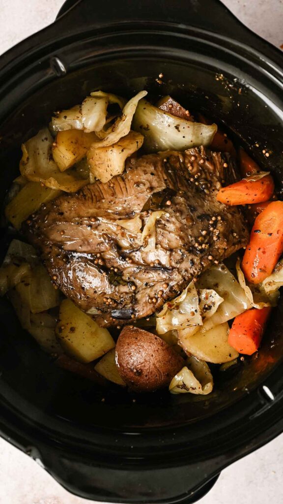 A delicious Slow Cooker Corned Beef with Cabbage sits alongside potatoes and carrots in a black slow cooker, ready to serve.