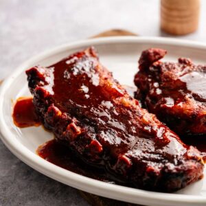 Two pieces of Slow Cooker Ribs coated in sauce are served on a white plate, placed on a wooden board.