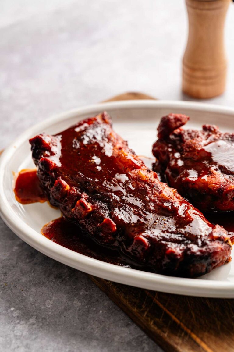 Two pieces of Slow Cooker Ribs coated in sauce are served on a white plate, placed on a wooden board.