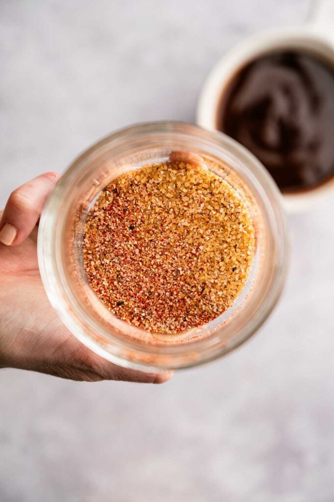 A hand holds a glass jar filled with a mixed seasoning blend for Slow Cooker Ribs; a blurred bowl of sauce is visible in the background.