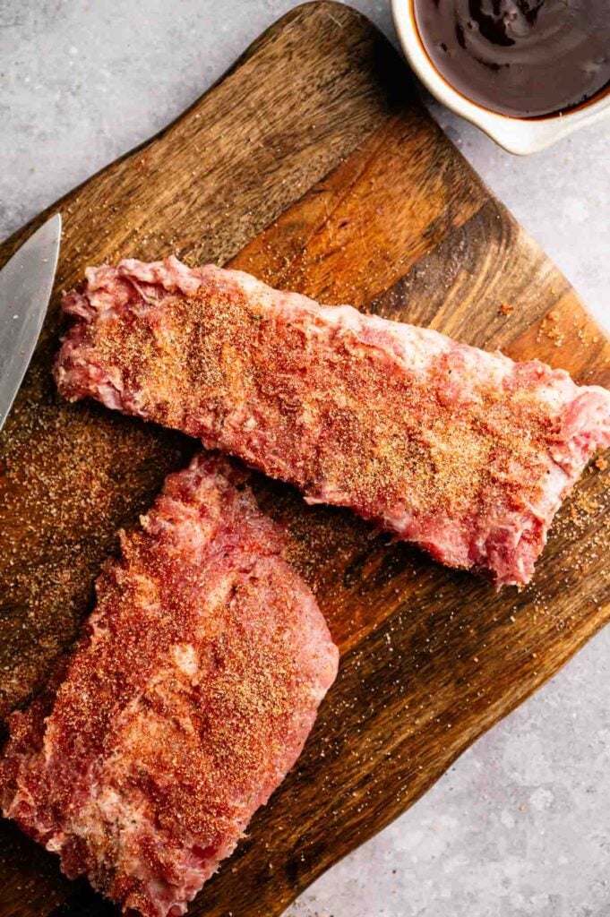 Two uncooked racks of ribs coated with dry seasoning sit on a wooden cutting board next to a knife and a small bowl of barbecue sauce—perfectly prepped for making tender Slow Cooker Ribs.