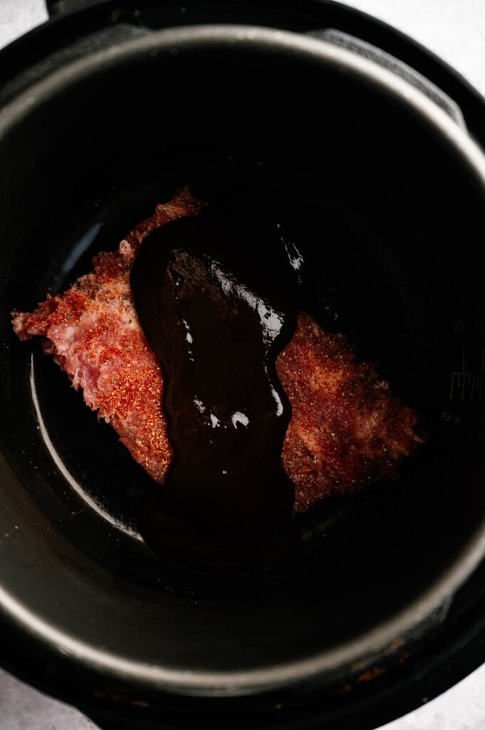 Raw seasoned meat with barbecue sauce on top sits at the bottom of a black slow cooker, ready to become tender and flavorful Slow Cooker Ribs.