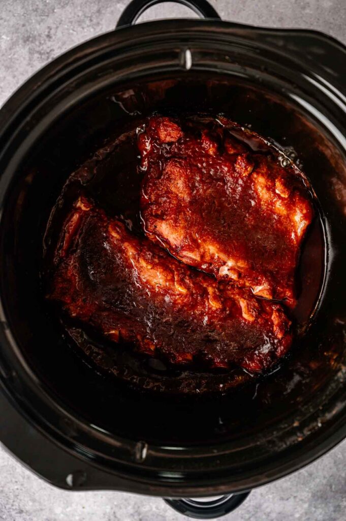 Two tender Slow Cooker Ribs covered in barbecue sauce rest inside a black slow cooker, ready to serve.