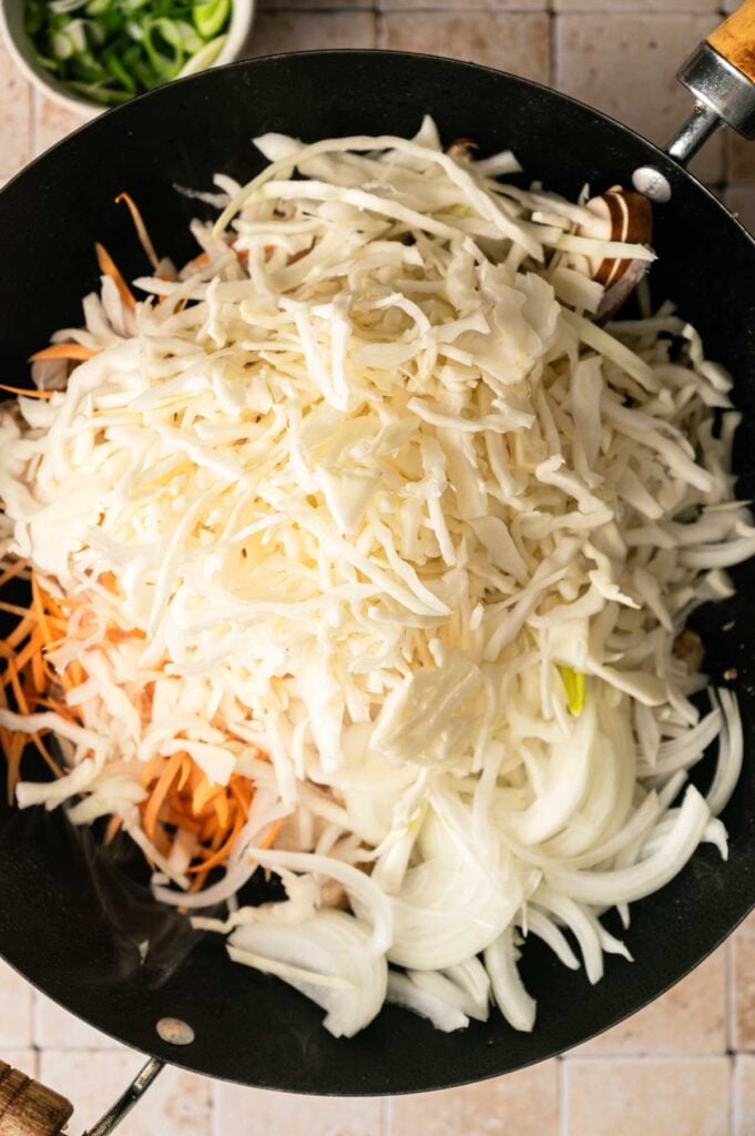 Shredded cabbage, sliced onions, and carrots in a black wok, ready to be cooked for yakisoba; a bowl of chopped green onions is visible in the background.