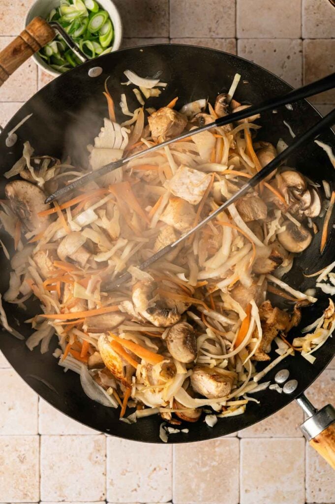 A wok filled with stir-fried vegetables, mushrooms, and tofu, reminiscent of savory yakisoba, being tossed with chopsticks; a small bowl of chopped green onions is nearby.