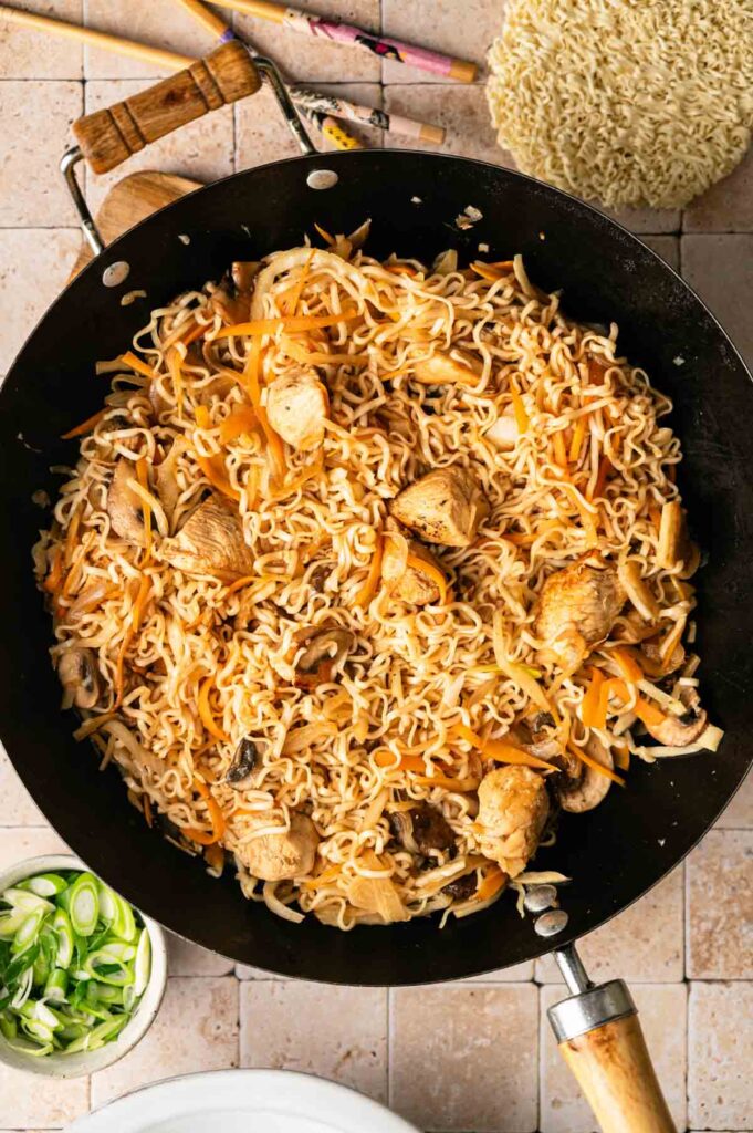 A wok filled with cooked yakisoba noodles, chicken pieces, sliced carrots, mushrooms, and onions on a tiled surface.