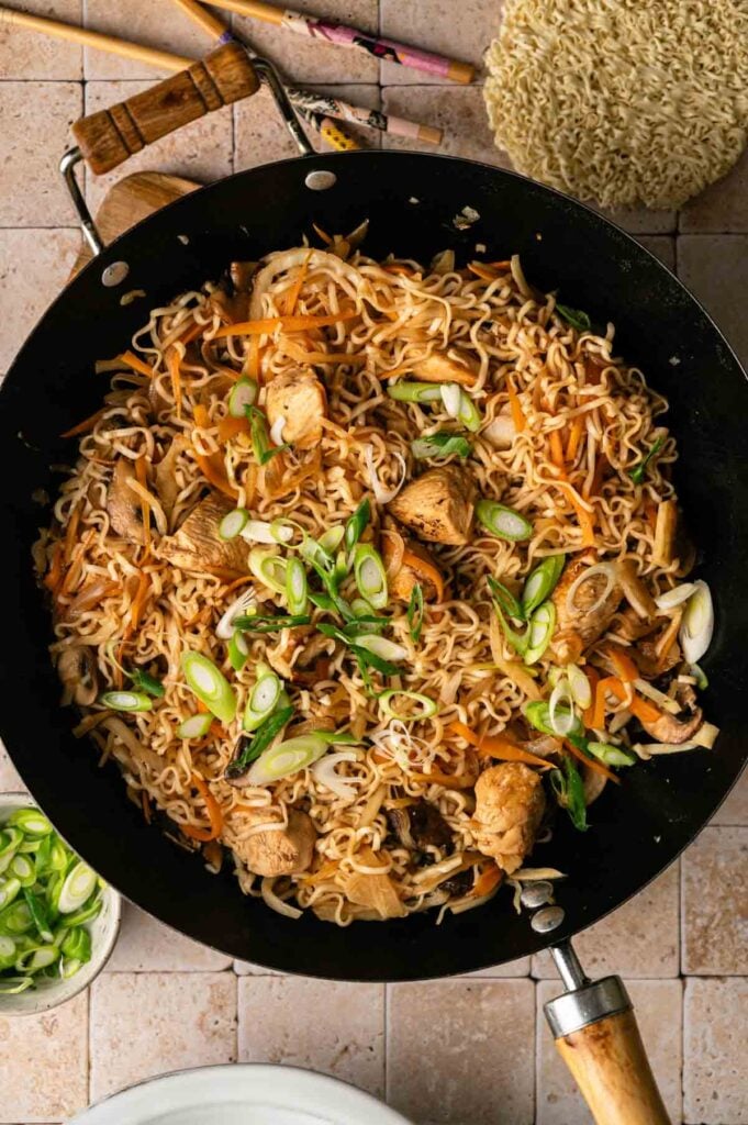 A wok filled with cooked ramen noodles, sliced chicken, carrots, mushrooms, and green onions—reminiscent of a hearty yakisoba dish—with chopsticks and uncooked ramen in the background.