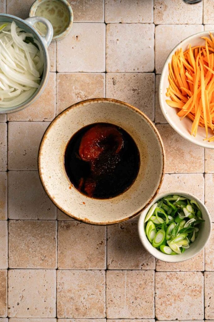 A bowl of dark yakisoba sauce sits in the center, surrounded by sliced onions, julienned carrots, and chopped green onions on a tiled surface.