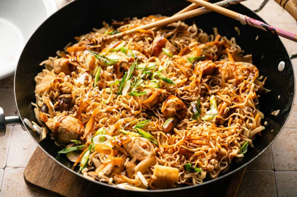 A pan filled with cooked yakisoba noodles, pieces of chicken, chopped green onions, and vegetables, with a pair of chopsticks resting on top.