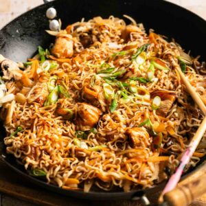 A wok filled with stir-fried yakisoba noodles, vegetables, and pieces of chicken, garnished with chopped green onions.
