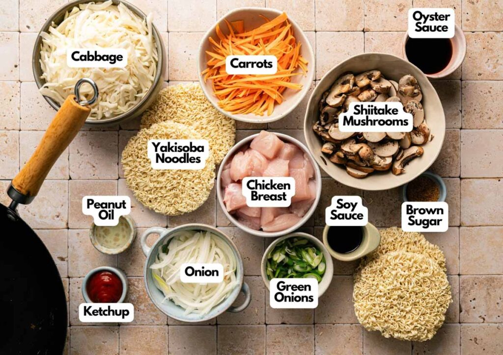 Bowls of sliced cabbage, carrots, shiitake mushrooms, chicken breast, yakisoba noodles, and various sauces and seasonings are arranged on a tiled surface, each labeled for easy yakisoba preparation.