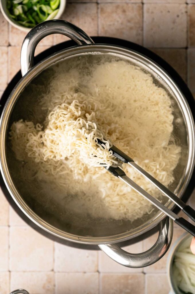 Tongs lifting cooked ramen noodles from a pot of boiling water, with bowls of green onions and yakisoba sauce nearby on a tiled surface.