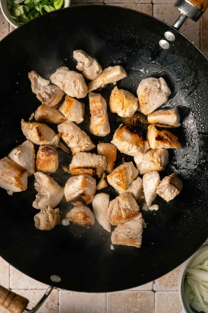 Chunks of cooked chicken breast are browning in a black wok on a stovetop, ready to be tossed with yakisoba noodles, with sliced green onions and onions visible in bowls nearby.