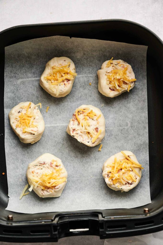 Six unbaked Air Fryer Chicken Pinwheels topped with shredded cheese are arranged on parchment paper inside an air fryer basket.