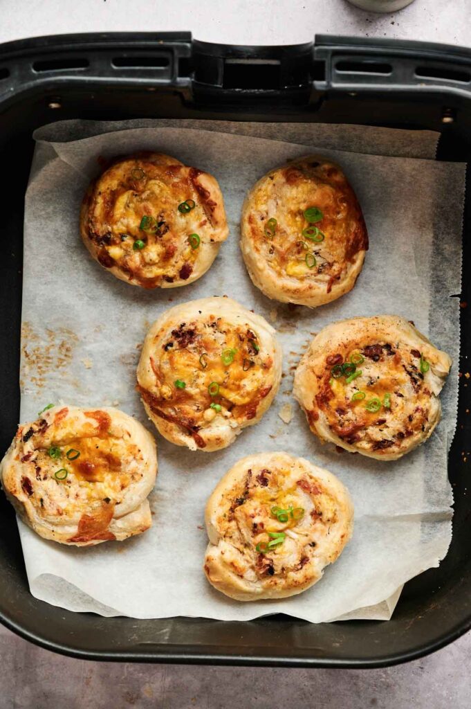 Six baked Air Fryer Chicken Pinwheels with cheese and bacon, topped with chopped green onions, arranged on parchment paper inside an air fryer basket.