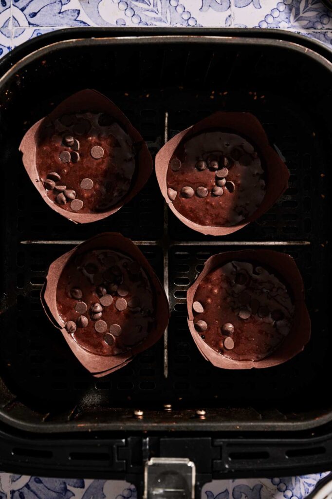 Four Air Fryer Chocolate Chip Muffins with chocolate chips in brown paper liners are placed inside an air fryer basket, ready to be baked.