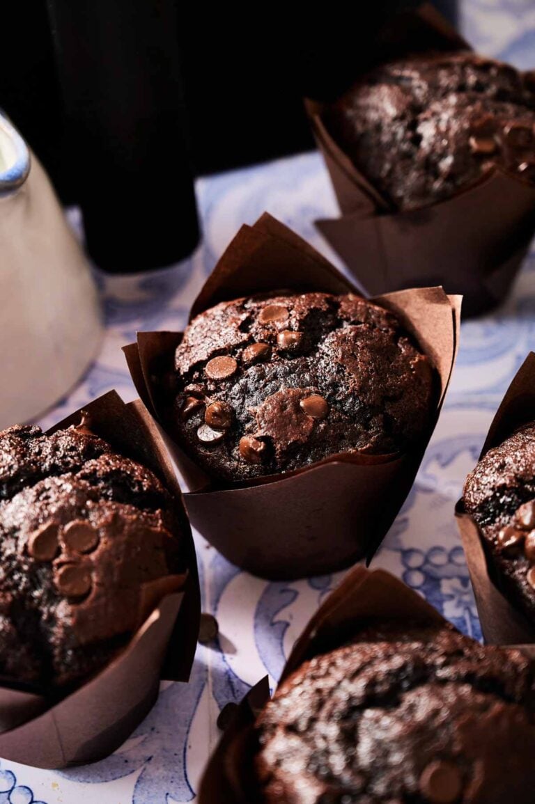 Five Air Fryer Chocolate Chip Muffins in brown paper wrappers are arranged closely together on a patterned surface, with chocolate chips visible on top of each muffin.