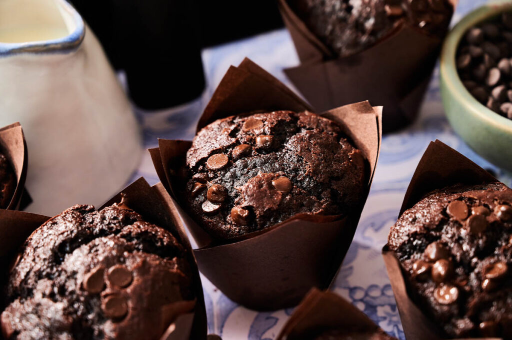 Close-up of Air Fryer Chocolate Chip Muffins in brown paper wrappers, with chocolate chips visible on top. A small pitcher and bowl of chocolate chips are nearby.