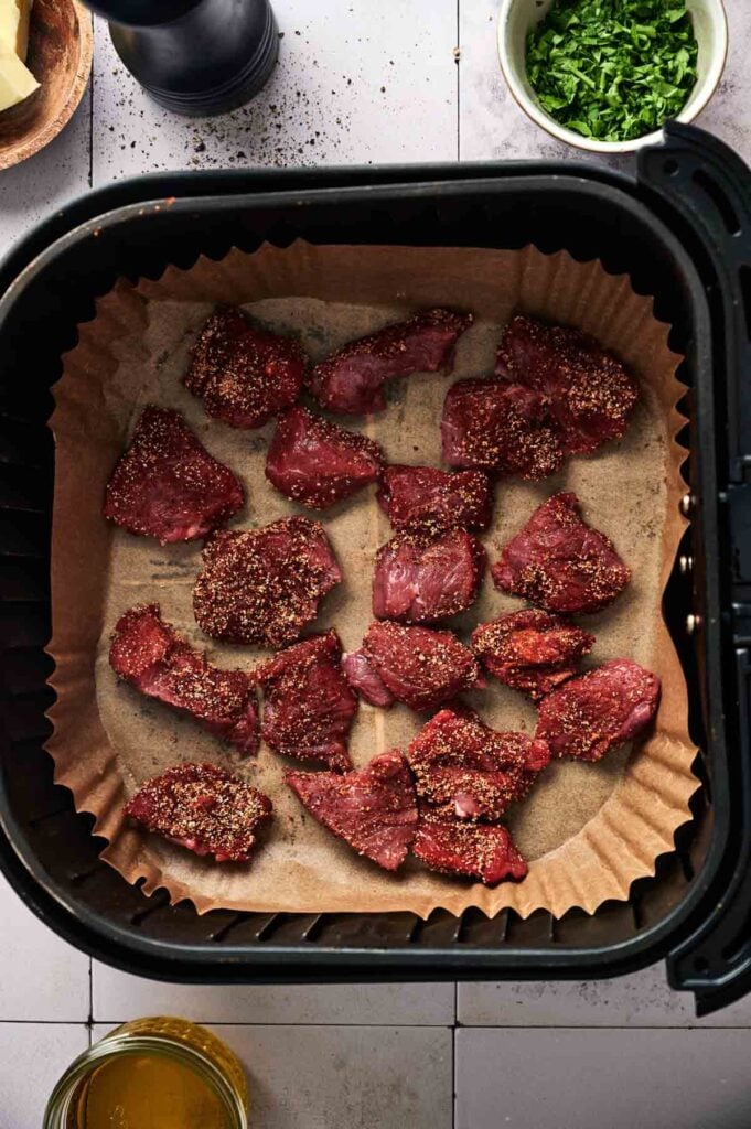 Raw, seasoned steak pieces are arranged on parchment paper in an air fryer basket, ready to be cooked for delicious Air Fryer Garlic Butter Steak Bites. Fresh herbs and other ingredients are visible in bowls nearby.