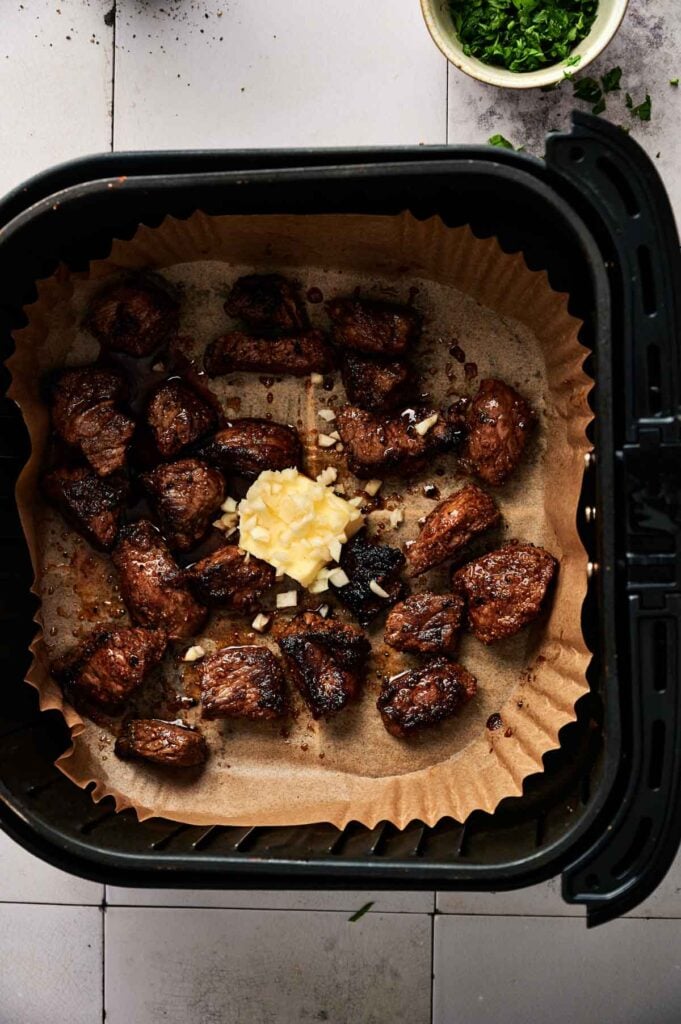 Air Fryer Garlic Butter Steak Bites cooked in a parchment-lined basket, topped with chopped garlic and butter, with fresh herbs visible nearby.