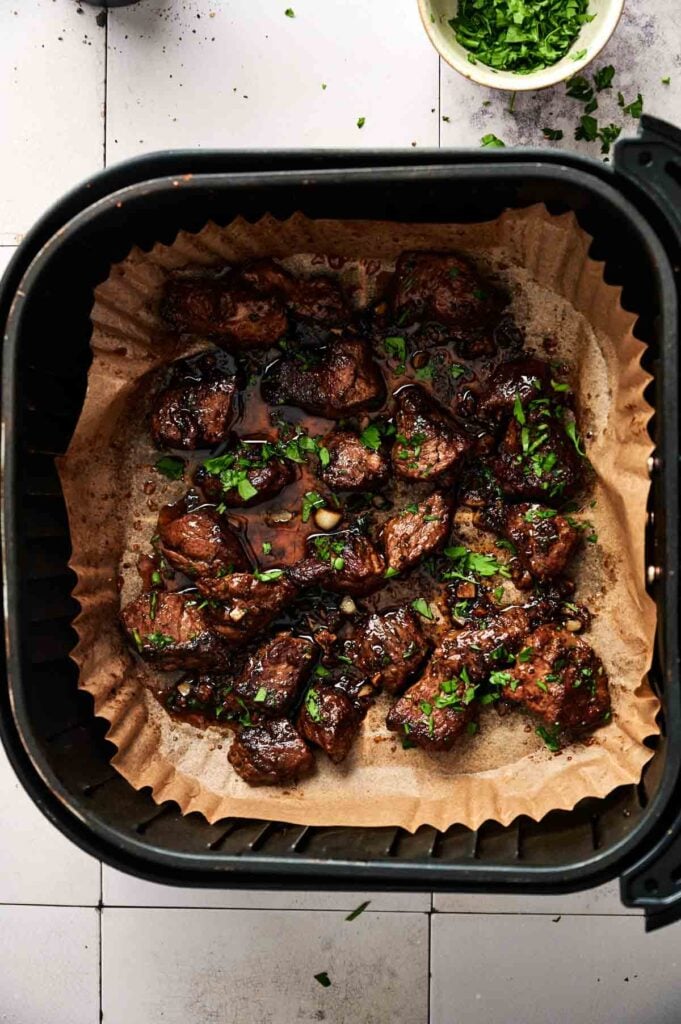 Air Fryer Garlic Butter Steak Bites with sauce and chopped herbs sit on parchment paper inside an air fryer basket, with a small bowl of herbs visible in the background.