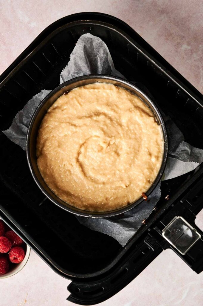 A round baking pan with uncooked Air Fryer Pineapple Upside Down Cake batter sits on parchment paper inside an air fryer basket; a small bowl of raspberries is nearby.