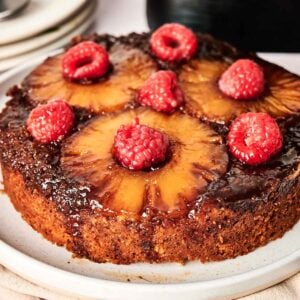 An Air Fryer Pineapple Upside Down Cake topped with whole pineapple rings and raspberries sits on a white plate.