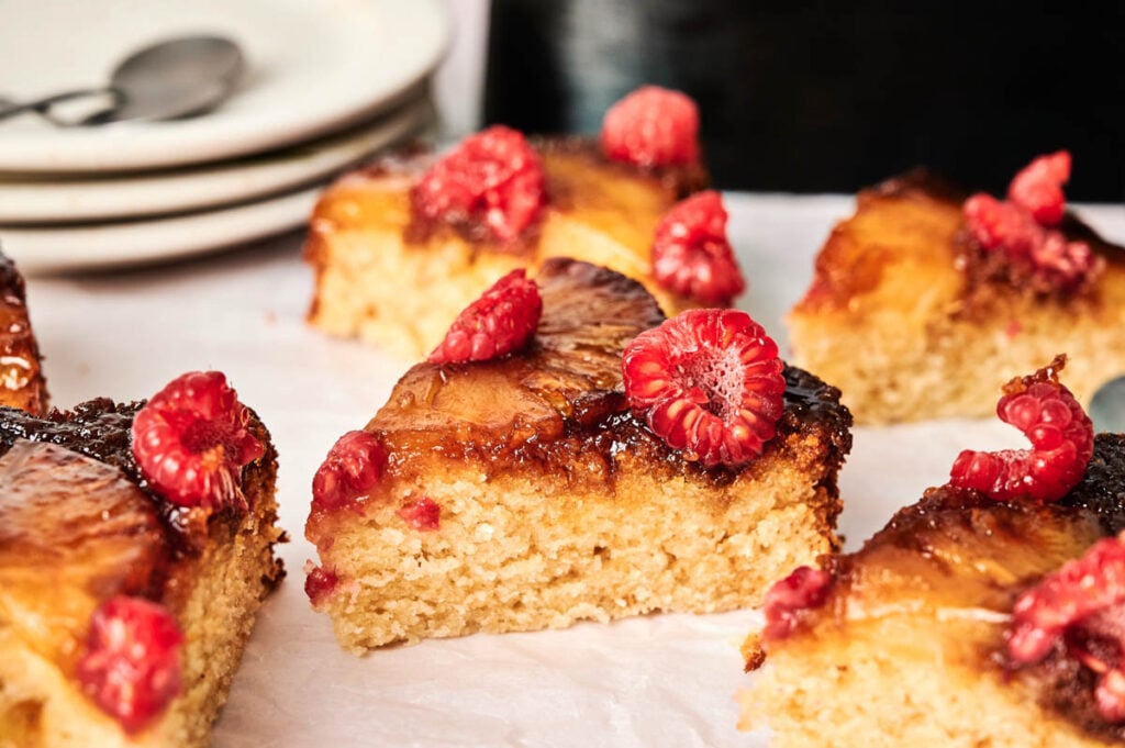 Sliced pieces of Air Fryer Pineapple Upside Down Cake topped with fresh raspberries are arranged on parchment paper, with plates and spoons in the background.