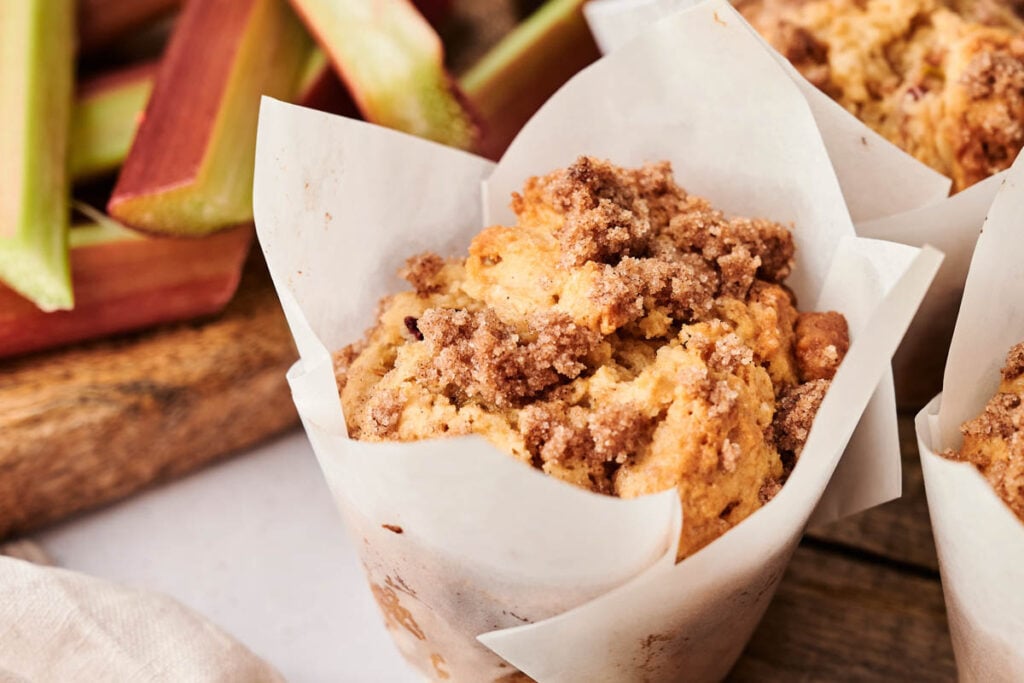 Close-up of Air Fryer Rhubarb Muffins with crumb topping in a white paper liner, with stalks of rhubarb blurred in the background.